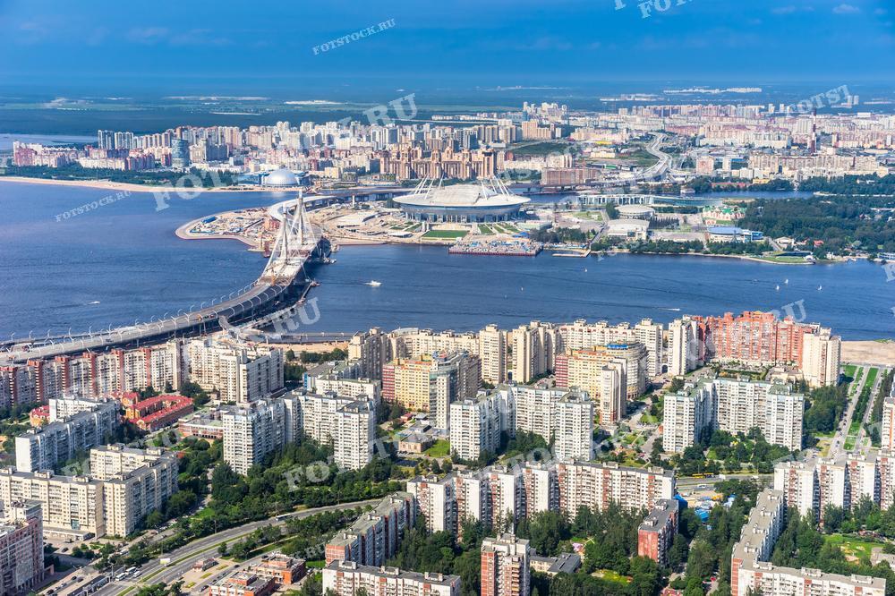 На фотографии представлен вид сверху на современный Санкт-Петербург. В центре изображения расположен мост через реку Нева, который соединяет два берега города. За мостом виднеется стадион на Крестовском острове, известный своей уникальной крышей в форме космического диска. Вокруг стадиона раскинулся жилой район с многоэтажными домами. Яркое солнечное освещение и чистое голубое небо делают композицию живой и динамичной. Эта фотография может использоваться в материалах о градостроительстве, архитектуре, а также в туристических гидах и рекламе городской недвижимости.